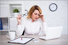 Middle-aged woman holding glasses while looking distraughtly at her computer.